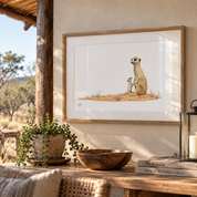 A framed art print featuring a watercolor illustration of two meerkats standing on a small patch of grass, displayed on a rustic wooden table in a warm, sunlit room setting.
