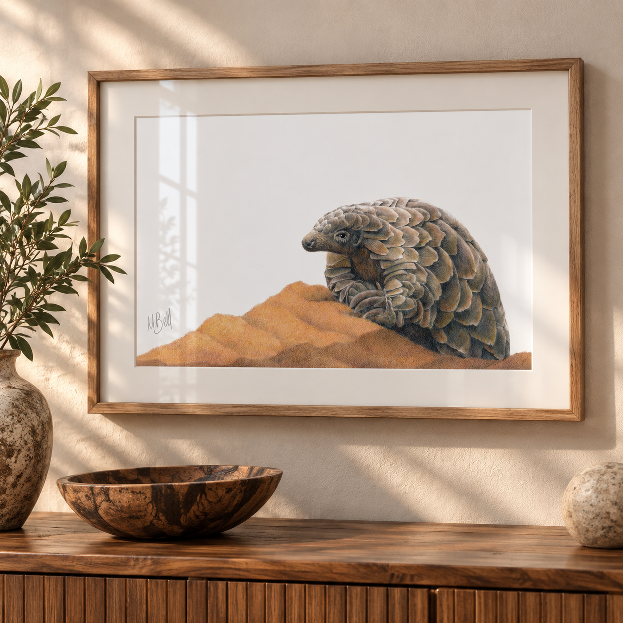 A framed art print of a pangolin resting on sand, displayed on a wooden console table in a styled room setting with a wooden bowl, a plant, and a stone sphere.