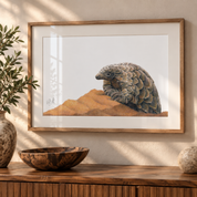 A framed art print of a pangolin resting on sand, displayed on a wooden console table in a styled room setting with a wooden bowl, a plant, and a stone sphere.