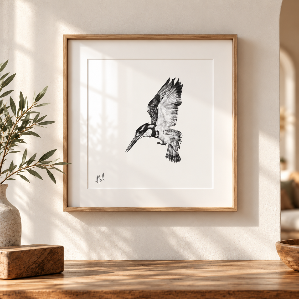 A framed black and white sketch of a flying Pied Kingfisher with a light wood frame and white mat, displayed on a wooden table against a warm beige wall with soft window shadows.