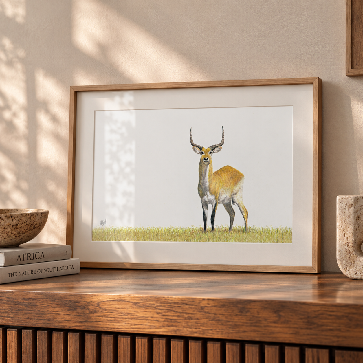 A framed art print of a red lechwe standing in the Okavango Delta, displayed on a wooden console table in a styled interior setting.