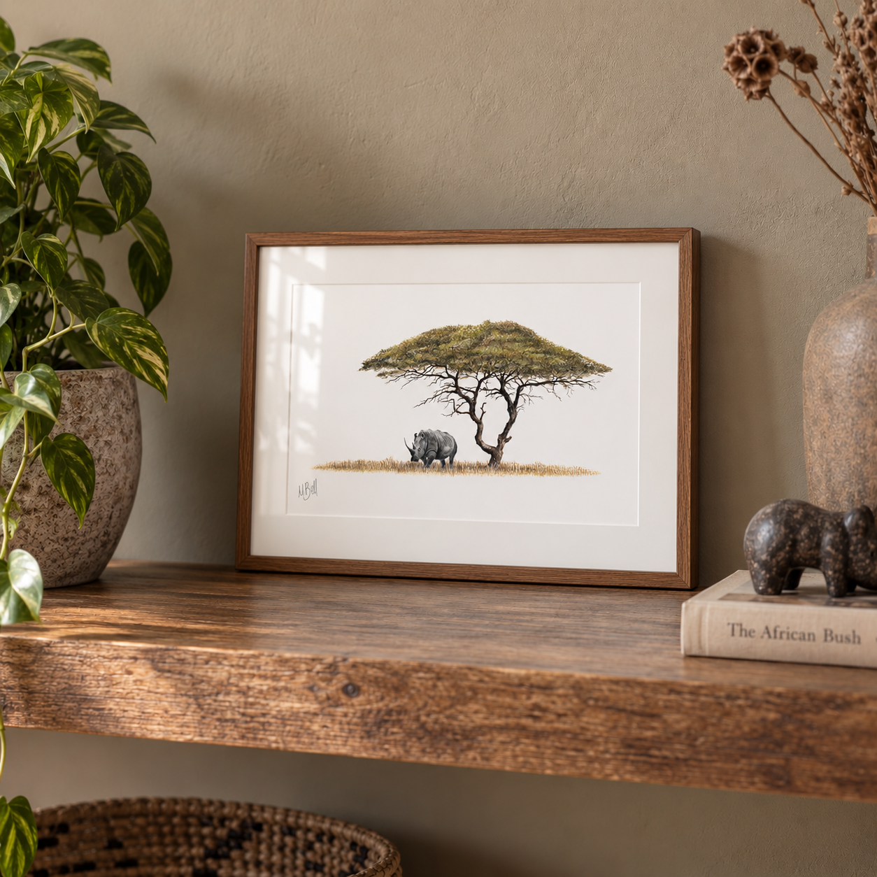 A framed art print depicting an acacia tree and a rhino, styled on a wooden shelf next to a potted plant and a decorative book.
