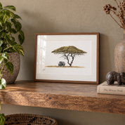 A framed art print depicting an acacia tree and a rhino, styled on a wooden shelf next to a potted plant and a decorative book.