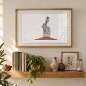 A framed art print of a scrub hare on a white background, hanging on a beige wall above a wooden shelf styled with books, a potted plant, ceramic vases, and a small botanical print.