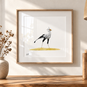 A framed art print featuring a watercolor illustration of a secretary bird standing on a yellow patch of grass, displayed on a wooden shelf against a warm beige wall with soft window light shadows.