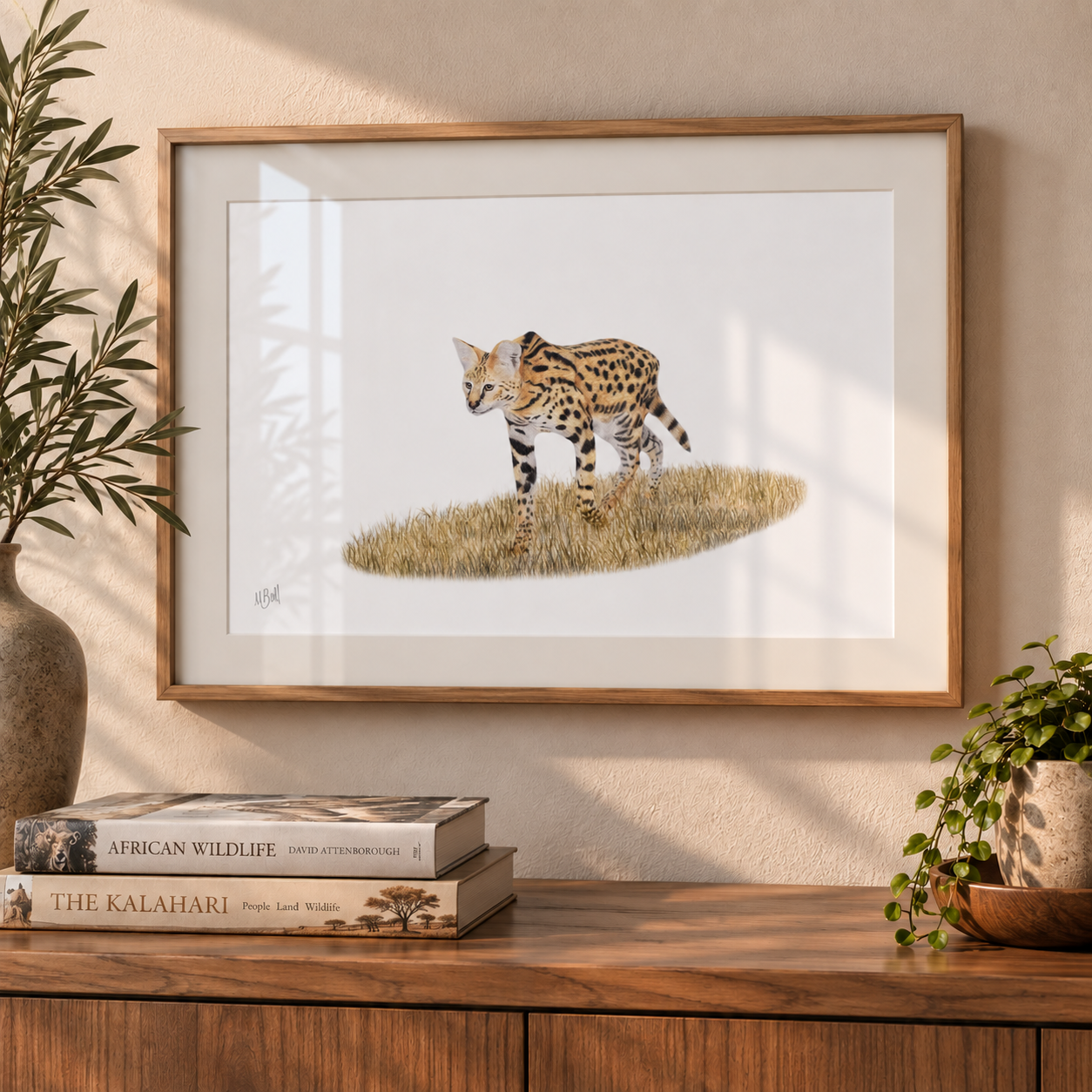 A framed art print of a serval cat standing on a patch of grass, displayed on a wooden console table in a styled room setting with books and potted plants.