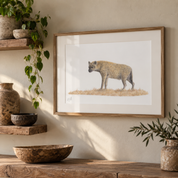 A framed art print featuring a detailed illustration of a spotted hyena standing on dry grass, displayed on a rustic wooden shelf in a warm, styled interior setting with plants and pottery.
