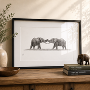A framed black and white pencil-style print of two elephants facing each other, displayed on a wooden console table with a vase, bowl, books, and a small elephant figurine in a styled interior setting.