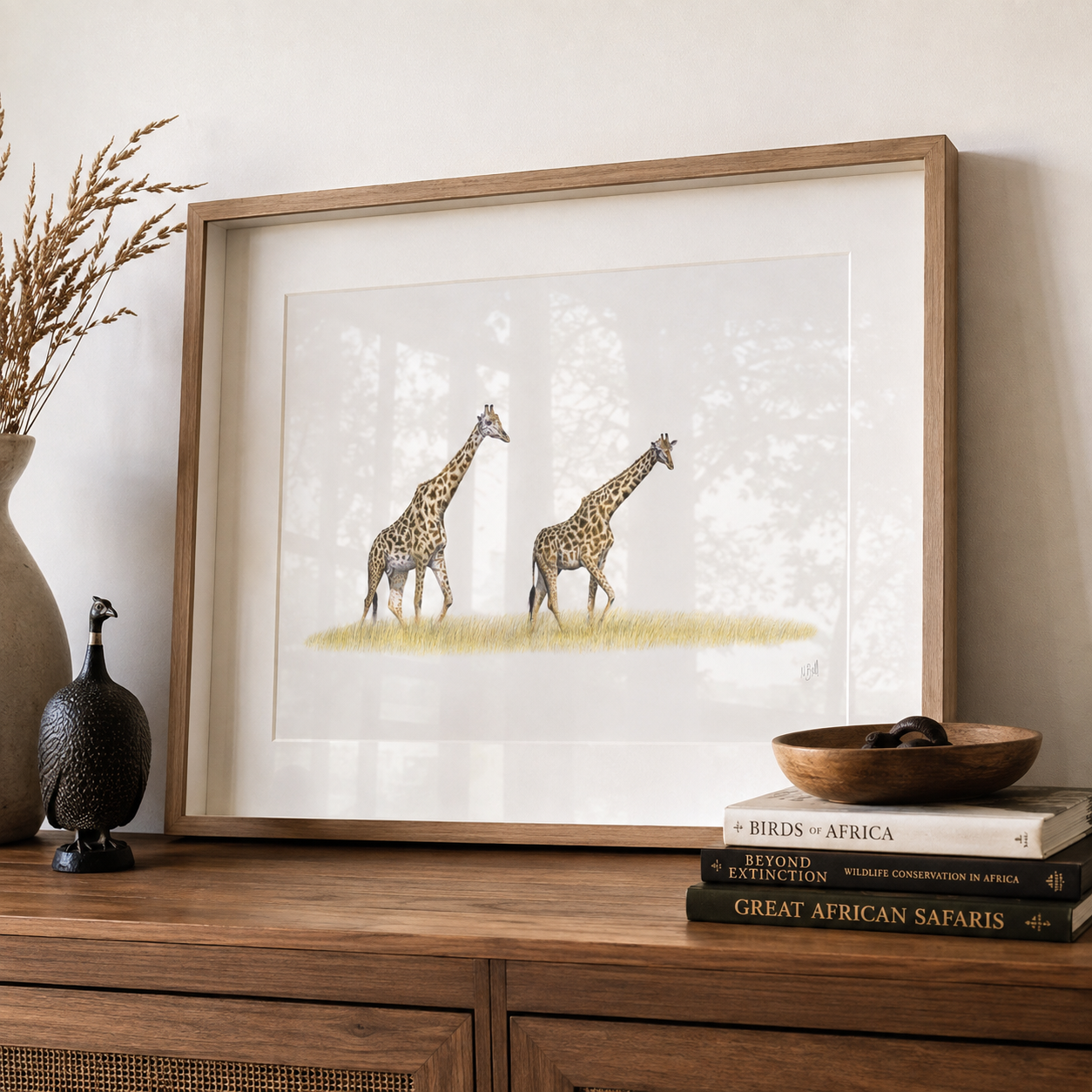 A framed art print featuring a watercolor-style illustration of two giraffes, displayed on a wooden console table with decorative books, a bowl, and a vase in a styled interior setting.