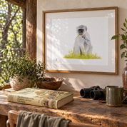 A framed art print of a gray vervet monkey sitting on green grass, displayed on a rustic wooden table in a warm, sunlit room with plants, a book, and binoculars.