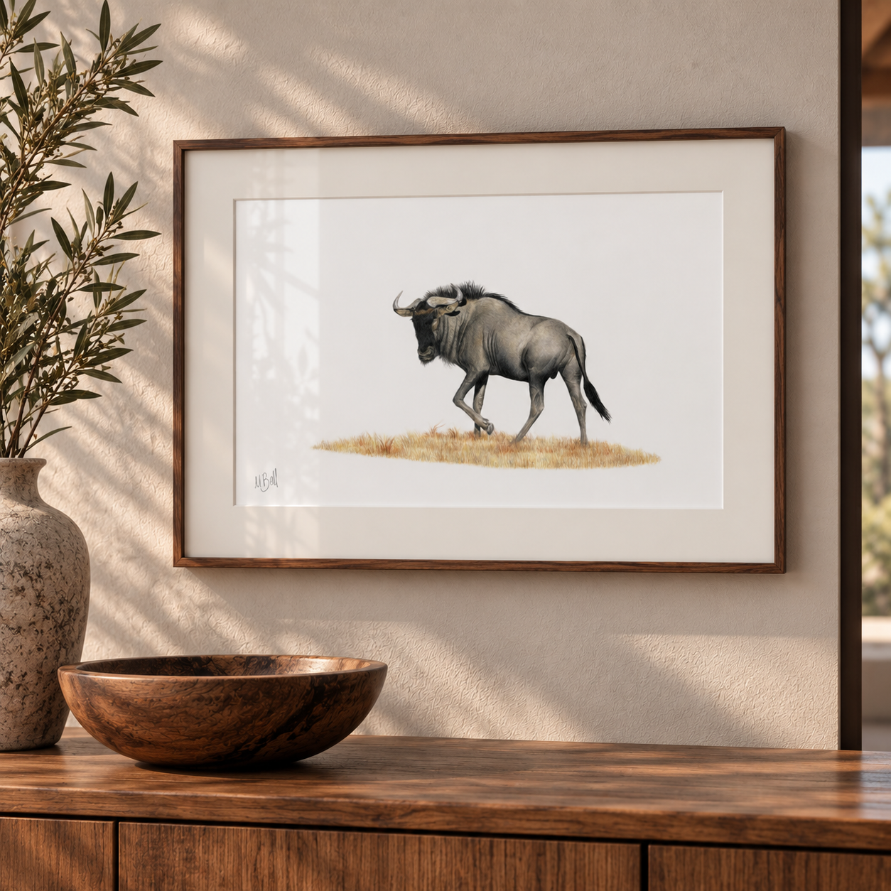 A framed art print of a wildebeest on a white background, styled on a wooden wall above a wooden console table with a bowl and vase.