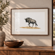A framed art print of a wildebeest on a white background, styled on a wooden wall above a wooden console table with a bowl and vase.