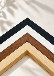 Four wooden picture frame corners in black, white, and natural wood tones arranged diagonally on a textured off-white wall with soft leaf shadows.