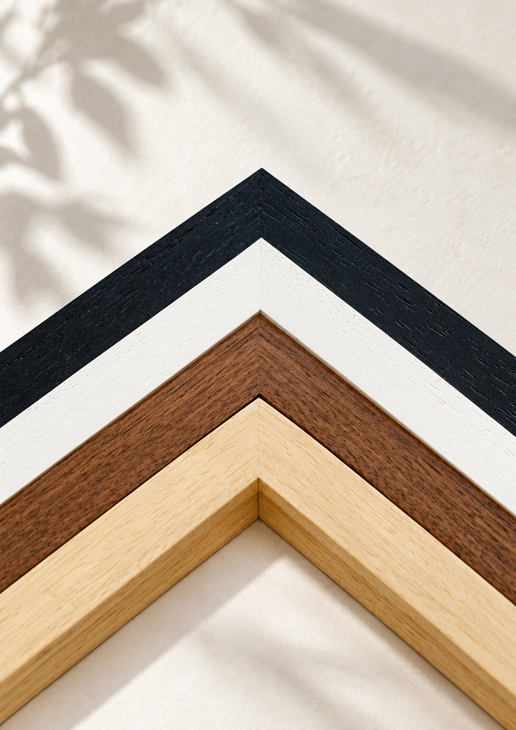 Four wooden picture frame corners in black, white, and natural wood tones arranged diagonally on a textured off-white wall with soft leaf shadows.