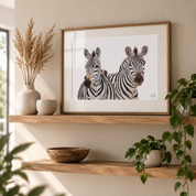 A framed art print depicting two zebras, styled on a wooden floating shelf in a warm, modern interior setting with dried grass in a vase and a green plant.